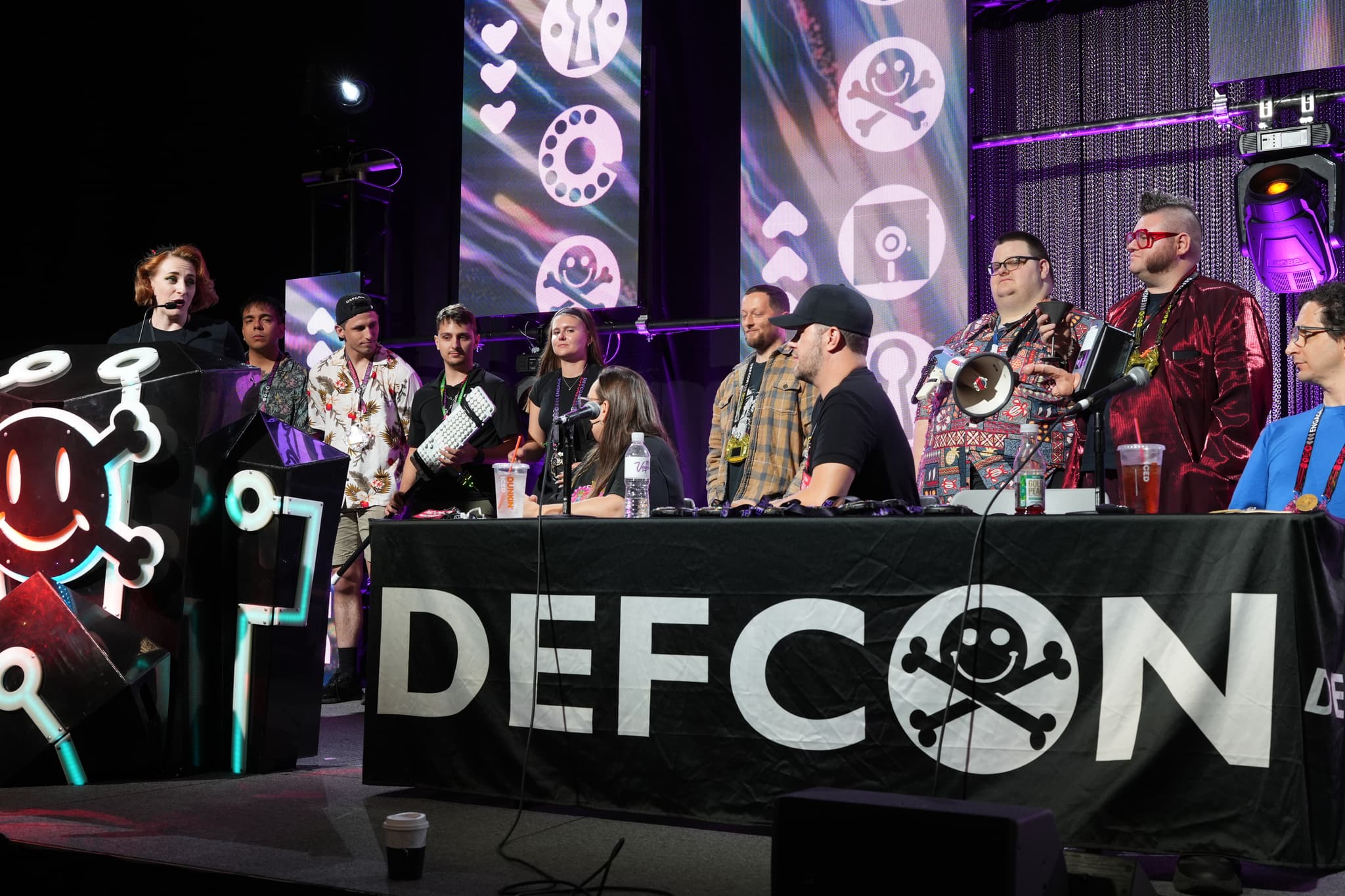 A group of people on a stage at DEFCON, with some standing and others seated behind a table, surrounded by technology-themed decorations and lighting