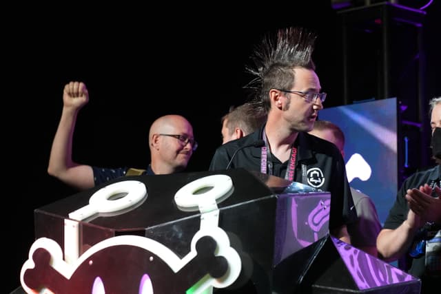 A group of people celebrating on stage, with one person raising a fist and another with a distinctive mohawk hairstyle standing behind a podium featuring a stylized skull design