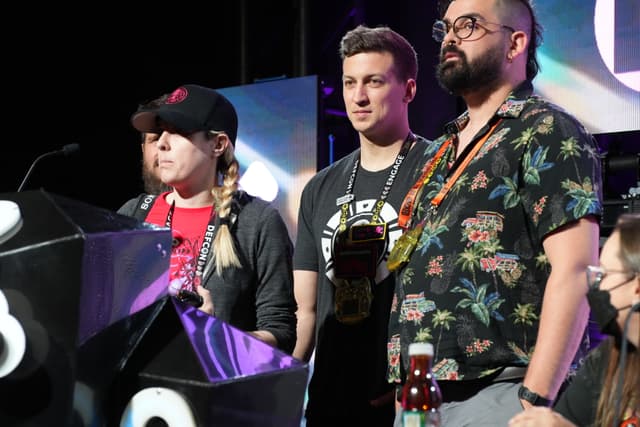 Three individuals standing on a stage, with one person speaking at a podium. The person at the podium is wearing a black cap and has blonde hair in braids. The other two individuals are standing behind, one wearing a black t-shirt and the other in a floral shirt. There is a microphone and some equipment visible on the podium