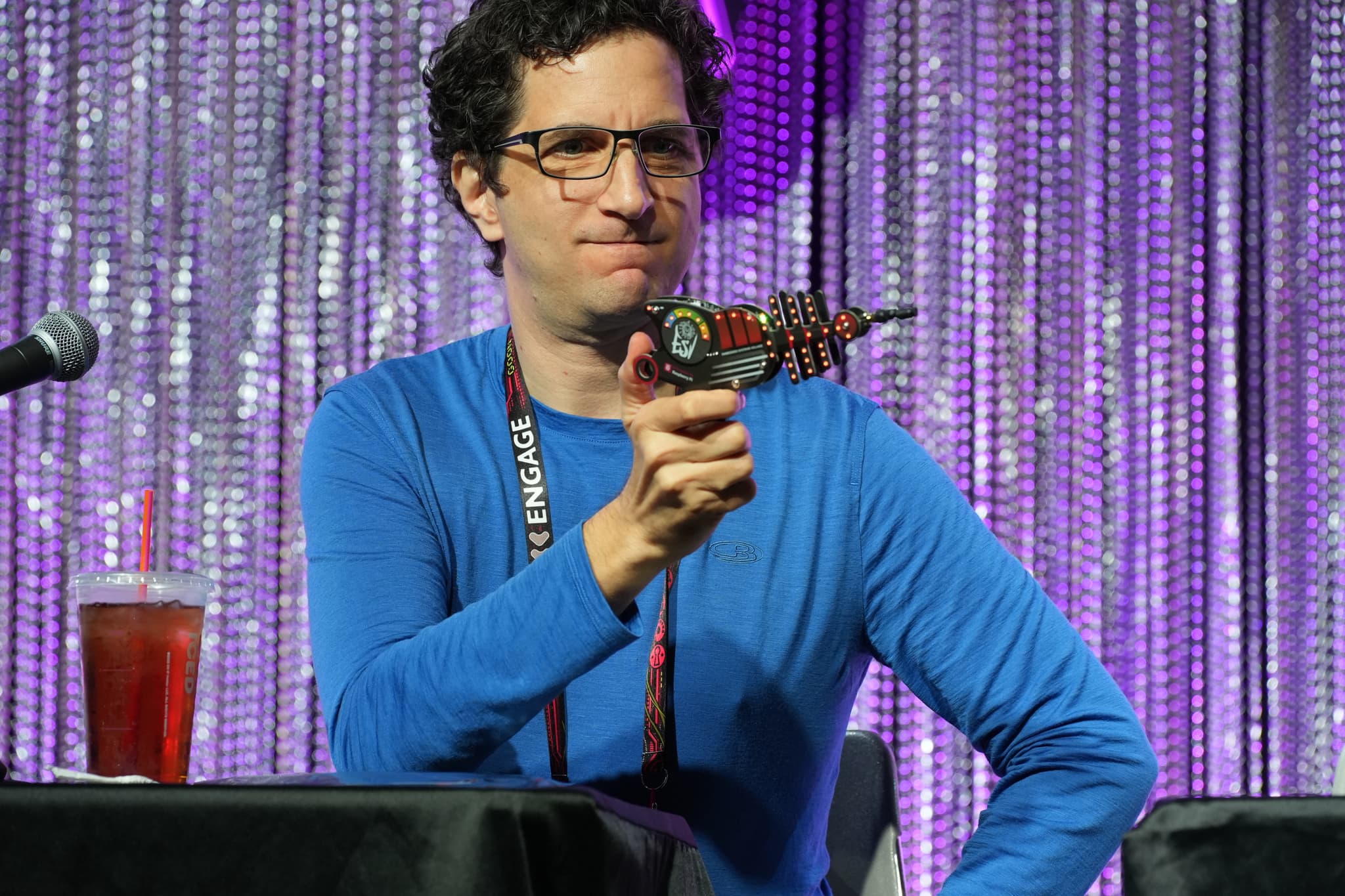 A man in a blue shirt holding a toy ray gun, sitting at a table with a microphone and a drink, in front of a sparkly curtain backdrop