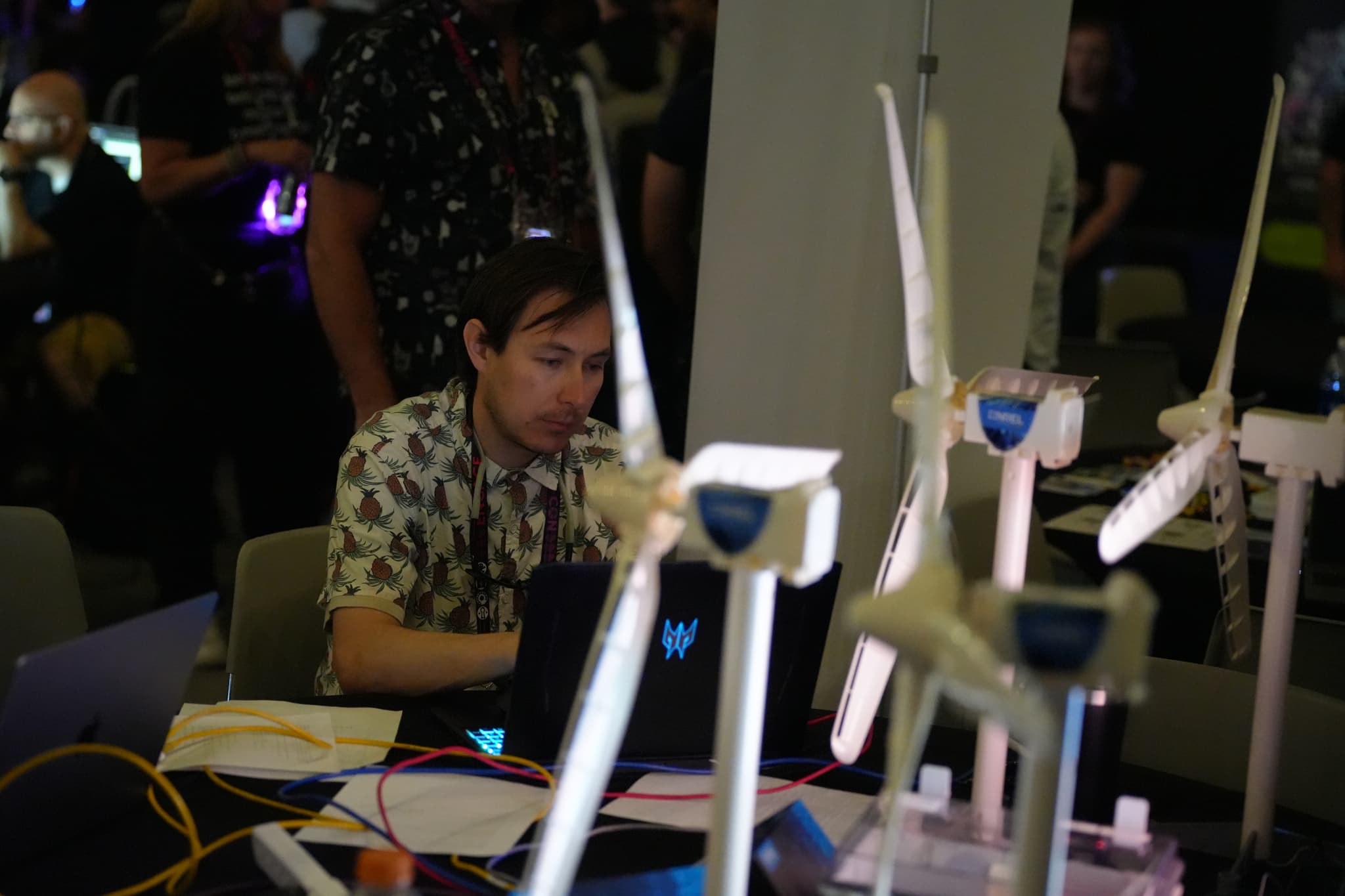 A person working on a laptop at a table with several small wind turbine models, surrounded by other people in a dimly lit environment