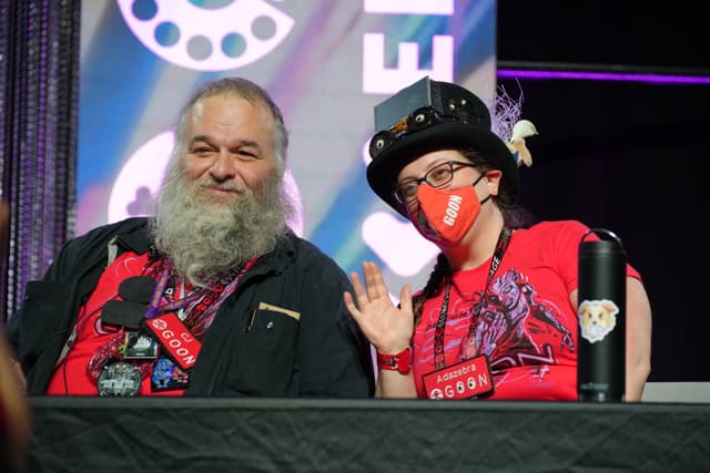 Two people sitting at a table, both wearing red shirts and badges. The person on the left has a long beard, while the person on the right is wearing a hat and a red face mask, waving at the camera. A colorful backdrop with text and designs is behind them