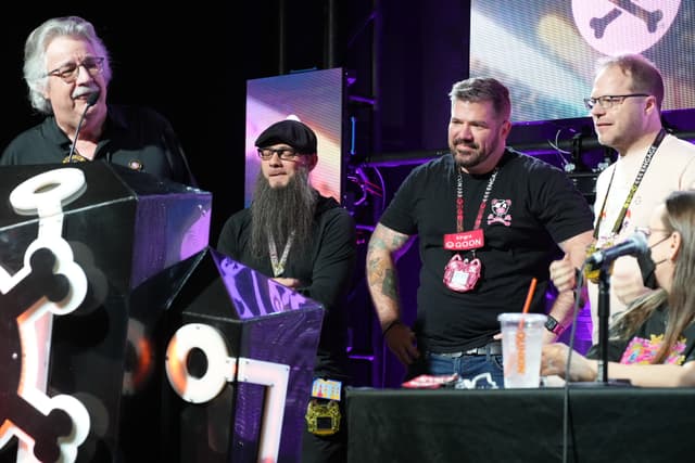 A group of people standing and sitting around a podium, engaged in conversation or presentation, with a backdrop featuring a large logo and stage lighting