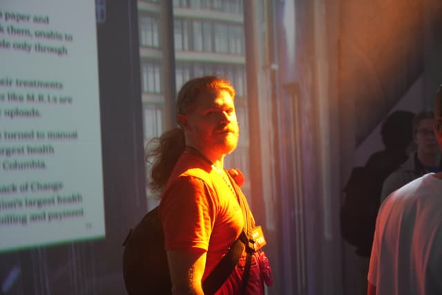 A person with a backpack stands in a room with sunlight streaming in, casting a warm glow. In the background, there is a projection of text and an image of a building. Other people are partially visible