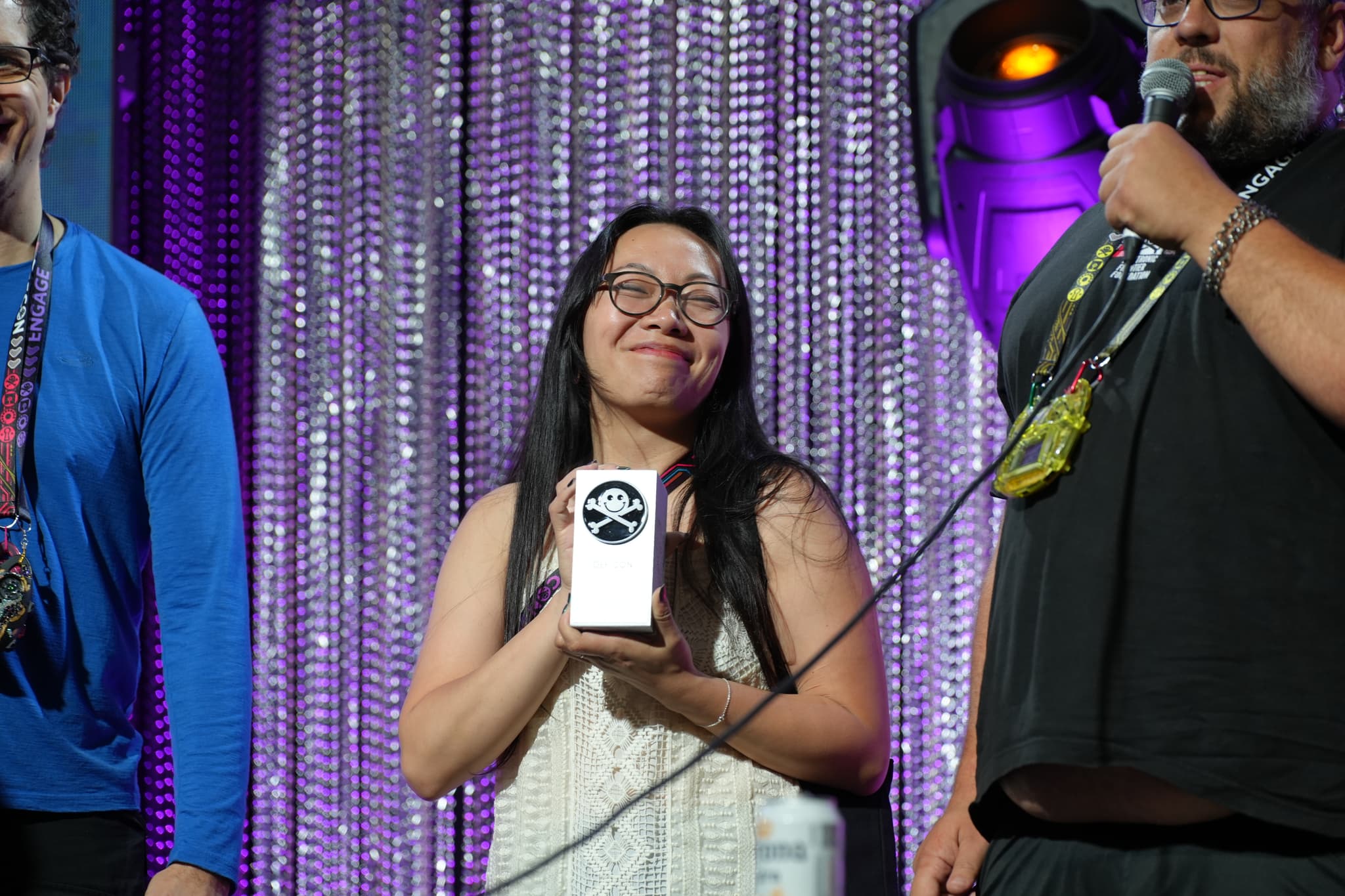 A person holding a trophy on stage, flanked by two other individuals, with a glittery curtain backdrop and stage lighting