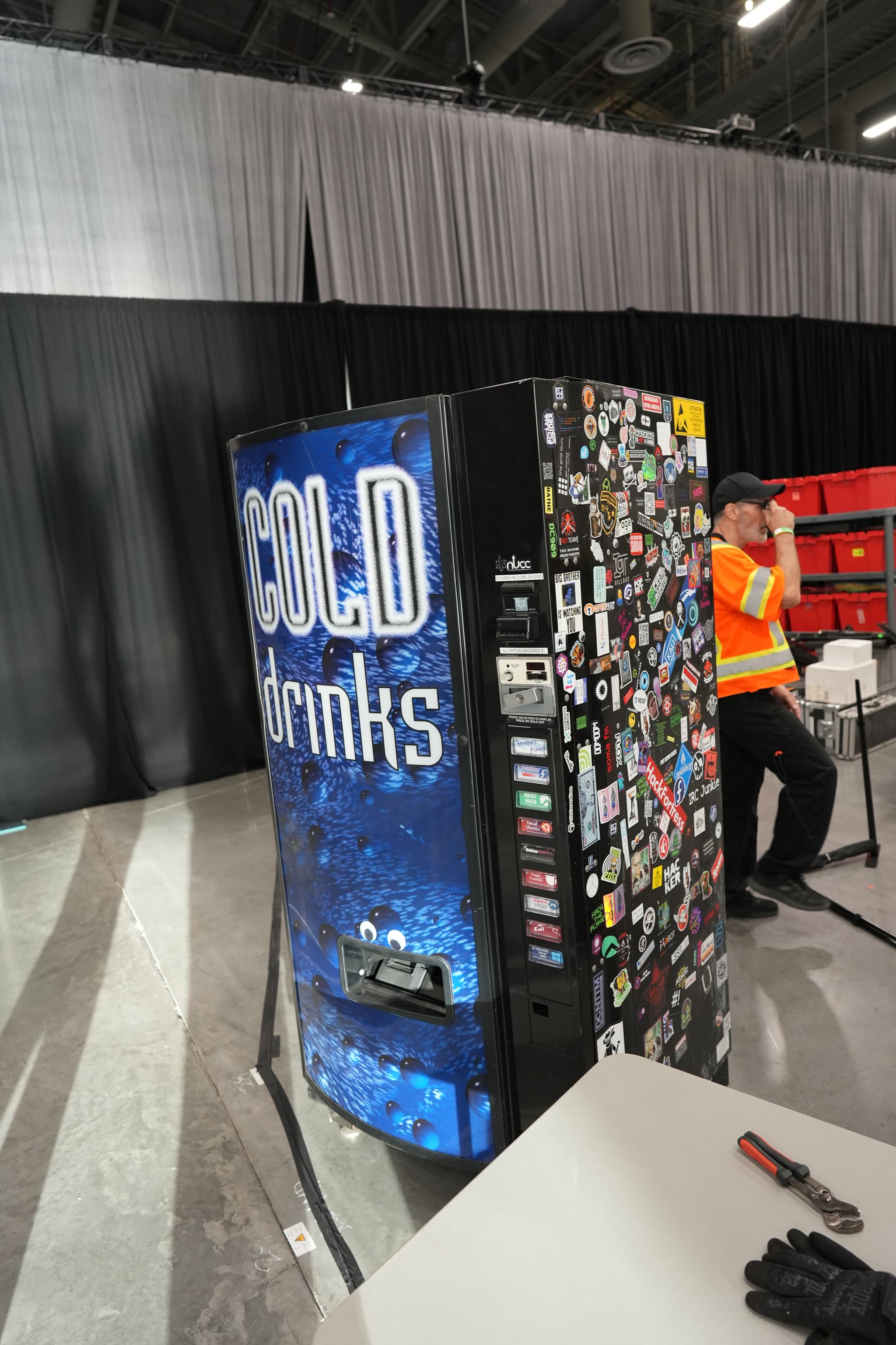 A vending machine labeled COLD drinks is situated in an indoor setting with a person in an orange shirt sitting nearby. The machine is covered with various stickers on one side, and there are tables and chairs around