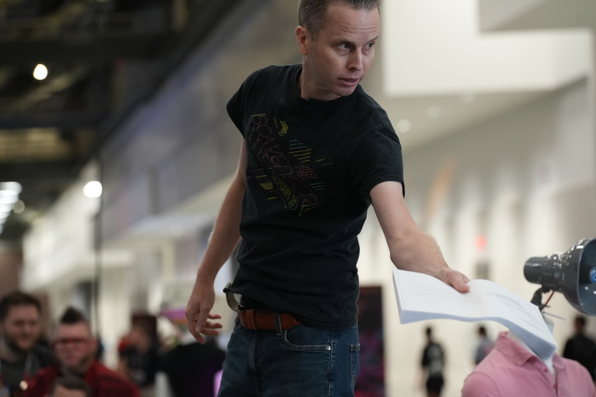 A person in a black t-shirt is extending a piece of paper towards a robotic arm in an indoor setting, with several people in the background