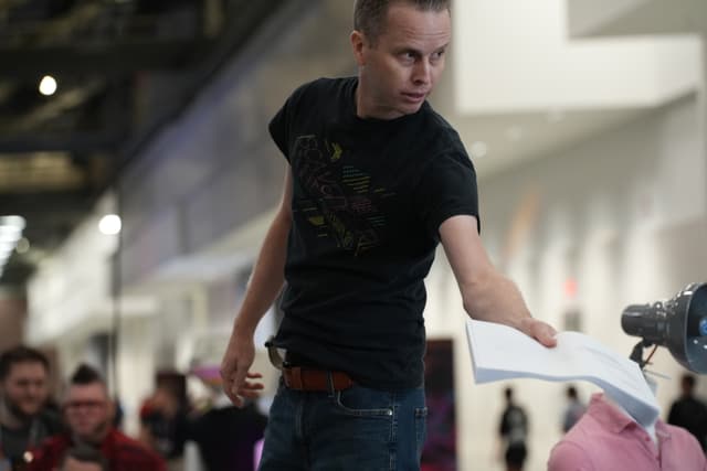 A person in a black t-shirt is extending a piece of paper towards a robotic arm in an indoor setting, with several people in the background