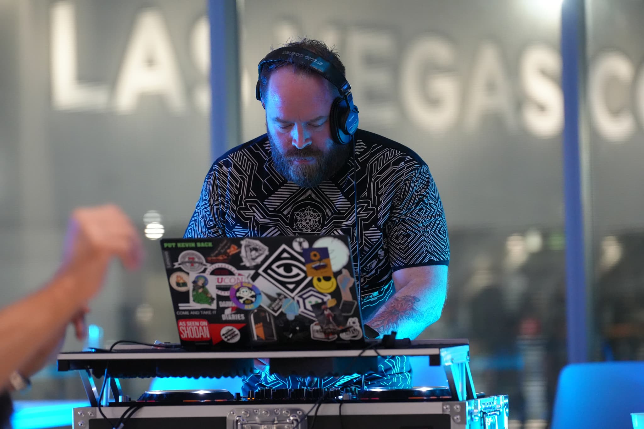 A DJ wearing headphones is performing at a setup with a laptop covered in various stickers. The background features large, blurred letters spelling LAS VEGAS