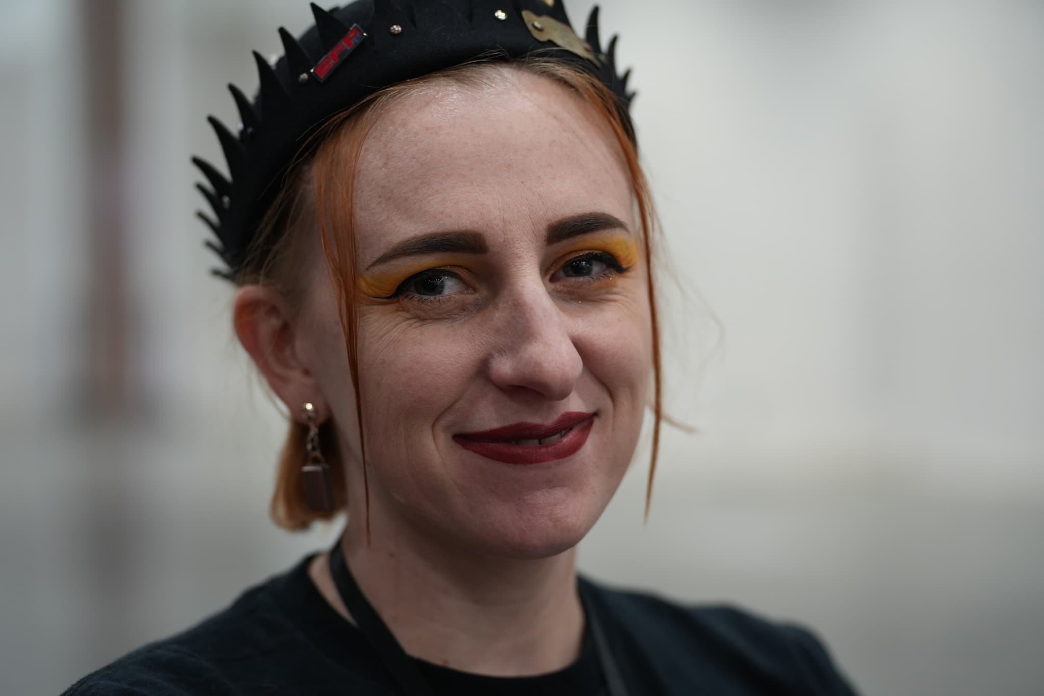 A person with light skin, wearing a black spiked headband, has orange hair and bold yellow eyeshadow, and is smiling slightly