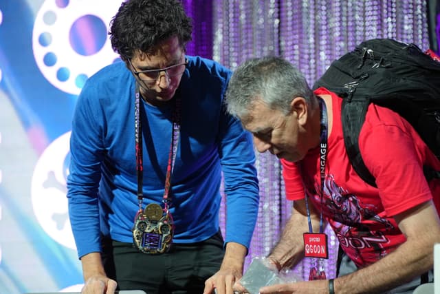 Two men are standing and looking down at something on a table, one wearing a blue shirt and the other a red shirt with a backpack. They are in a setting with a colorful, decorated background