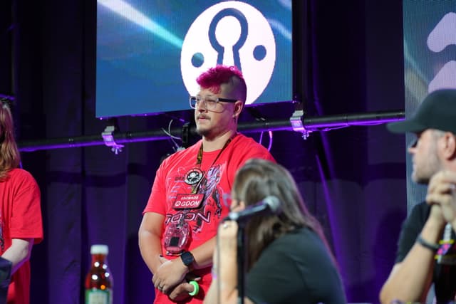 A group of people on a stage, with one person in the center wearing a red shirt and glasses, standing in front of a backdrop featuring a logo