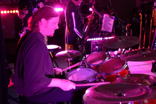 A drummer playing a drum set on stage, illuminated by pink and purple lights, with other musicians and equipment in the background