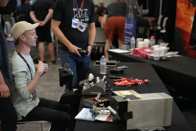 A person sitting at a table with various items, including a joystick and other equipment, while another person stands nearby. The background features more people and a booth setup with promotional materials and snacks