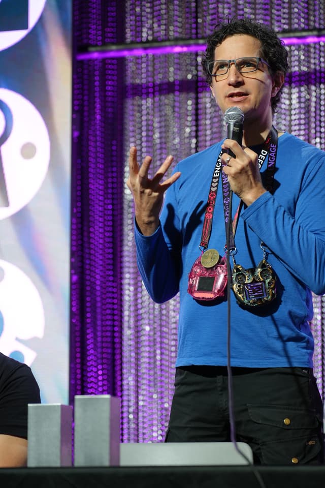 A person wearing a blue shirt and glasses is speaking into a microphone on a stage, with two medals hanging around their neck. The background features a purple curtain and part of a sign with large letters