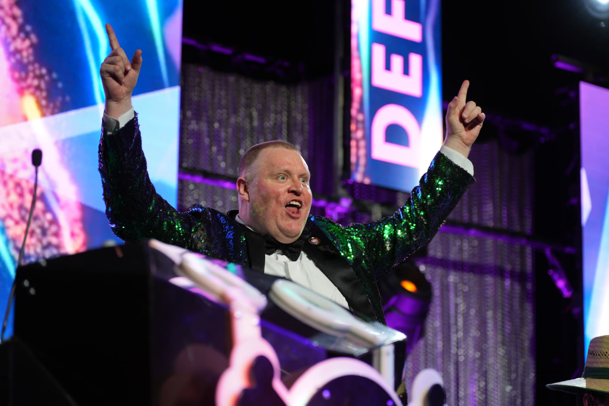 A man in a sparkly jacket and bow tie is standing with his arms raised, pointing upwards, and smiling enthusiastically on a stage with colorful lights and decorations