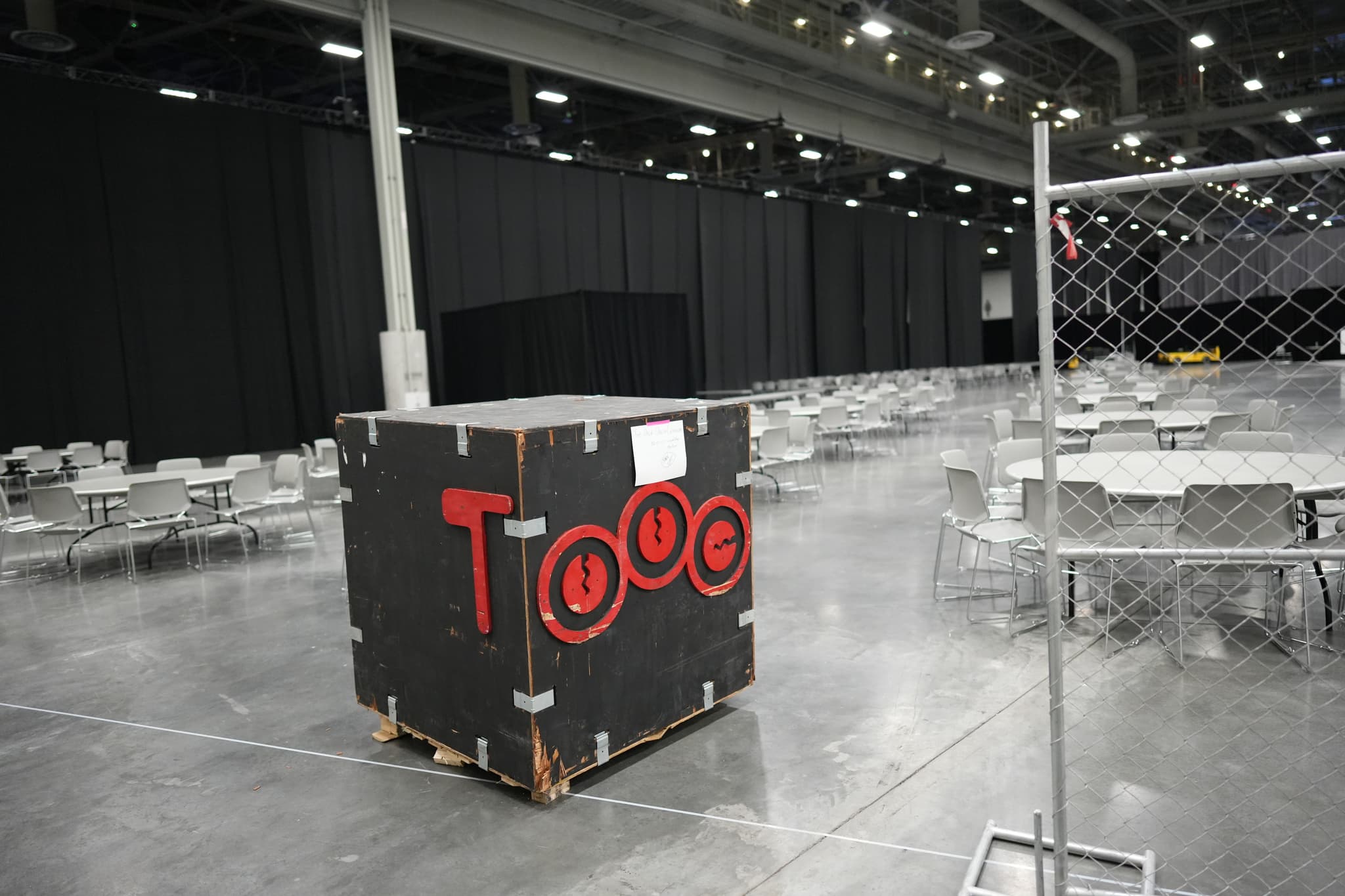 A large, mostly empty indoor space with a black crate labeled TOOC in red letters, surrounded by round tables and chairs, and a chain-link fence section on the right