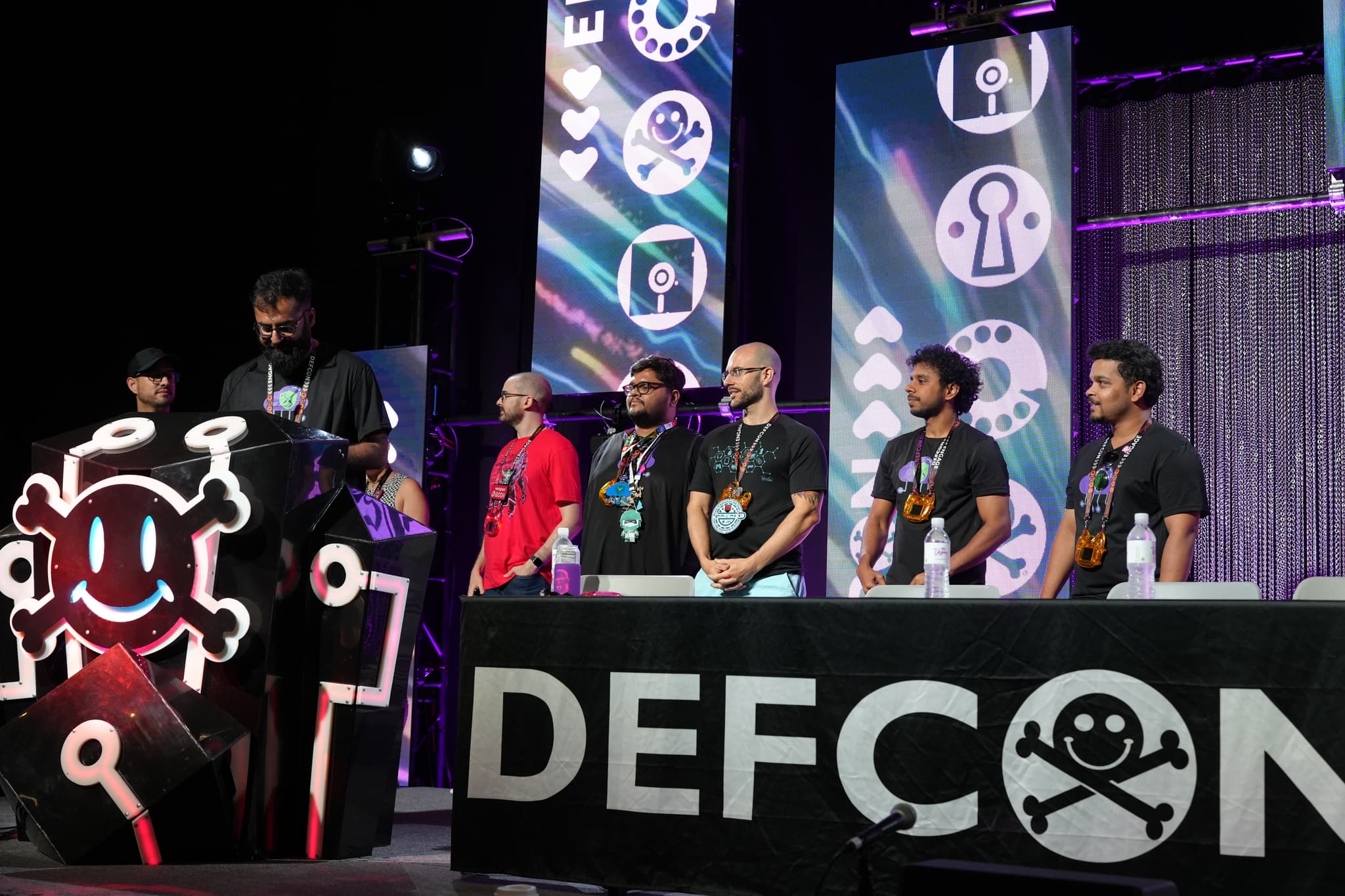 A group of people standing on a stage with DEFCON written on the front, featuring various tech-related symbols in the background