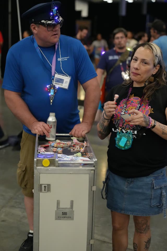 A man in a blue shirt and hat stands next to a woman in a black shirt and denim skirt. They are indoors, and the man is leaning on a cart with various items on top. The woman appears to be speaking or explaining something