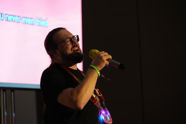 A person with a beard and glasses is singing into a microphone, wearing a black shirt and a lanyard, with a screen displaying text in the background