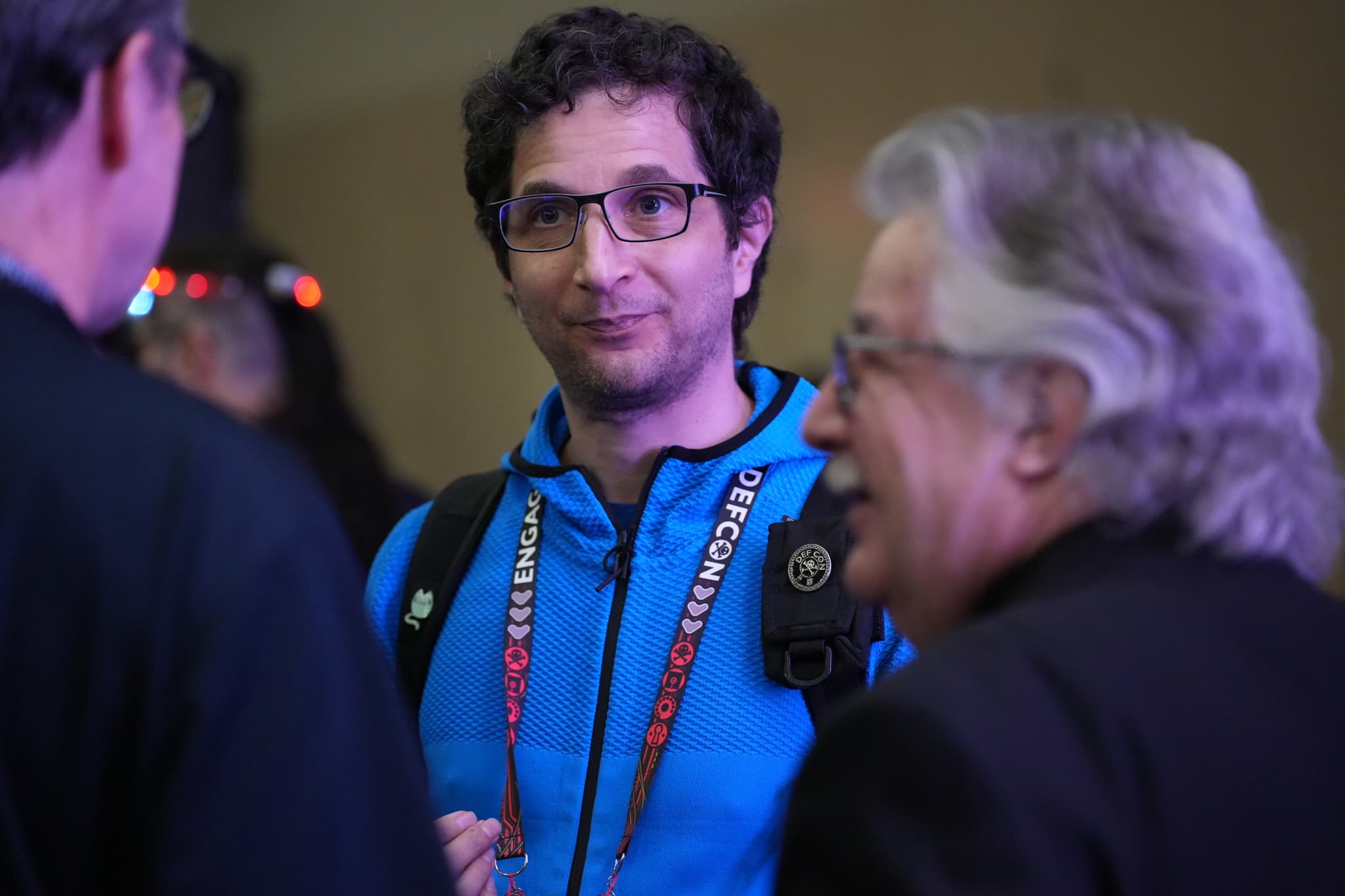 Three people engaged in conversation, with one person in the center wearing a blue jacket and glasses, and the other two partially visible from behind