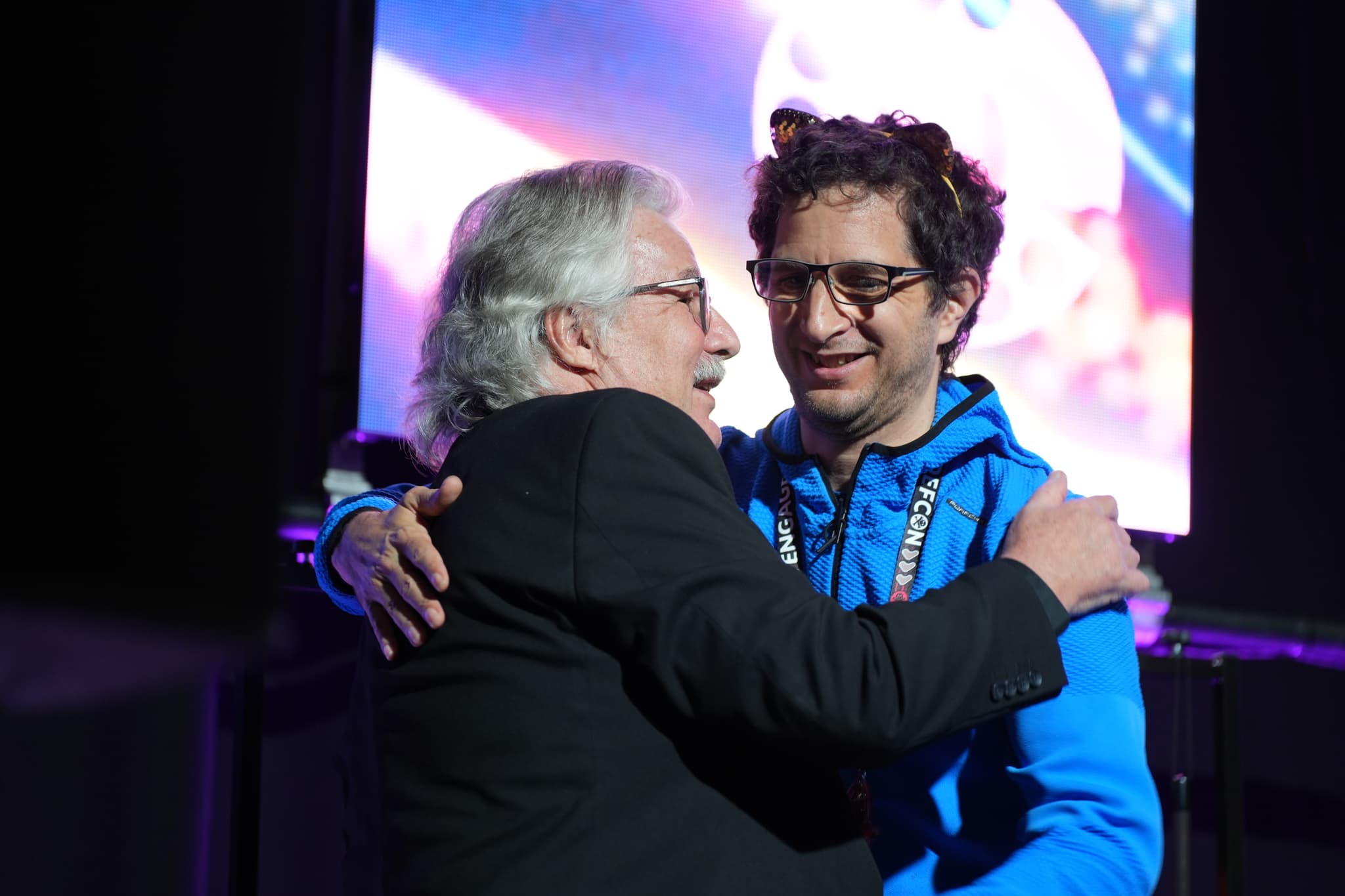 Two men embracing each other, one wearing a black suit and the other in a blue jacket, with a colorful background behind them