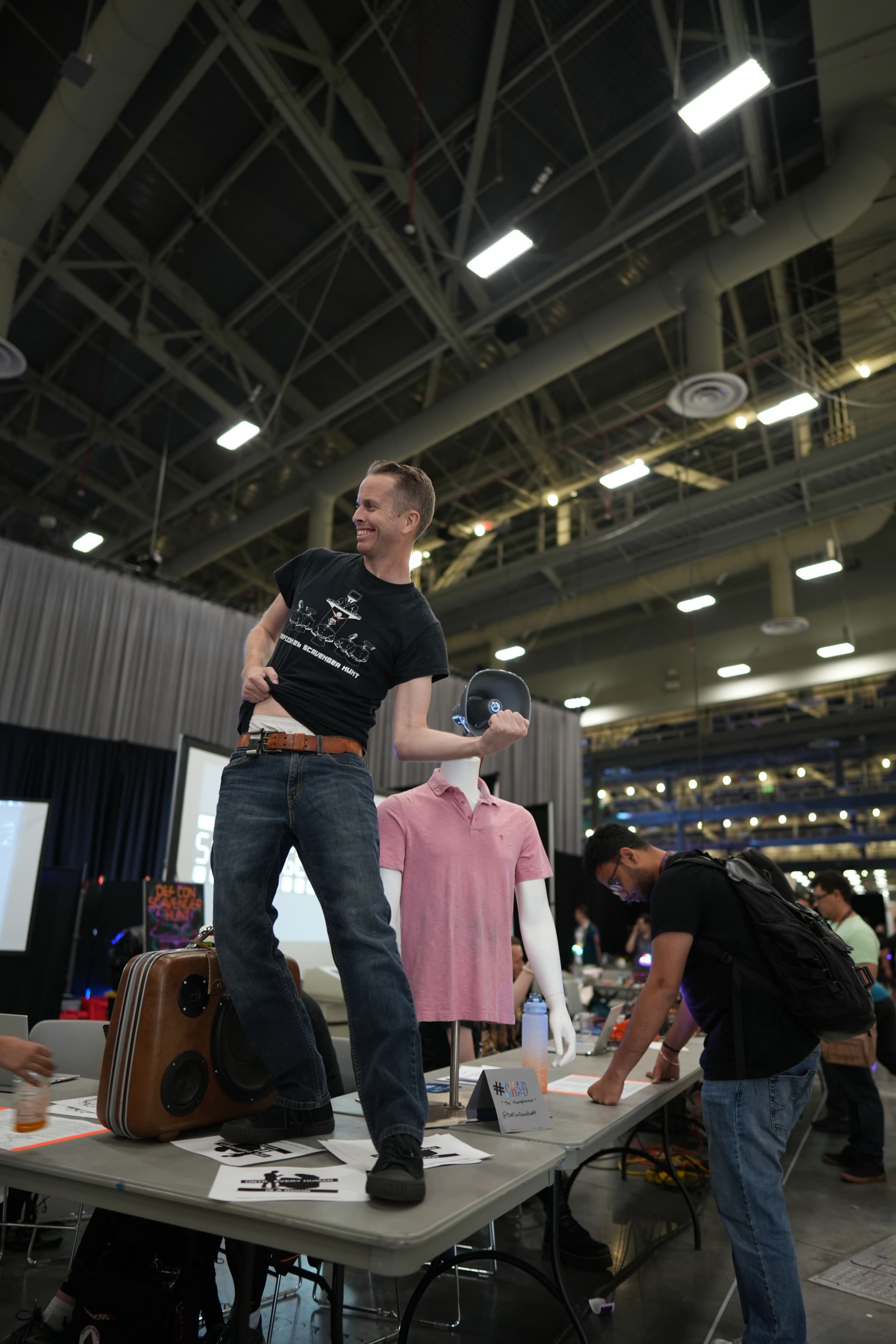 A person standing on a table in a large indoor venue, striking a pose with one arm raised and the other on their hip, while another person is leaning over the table