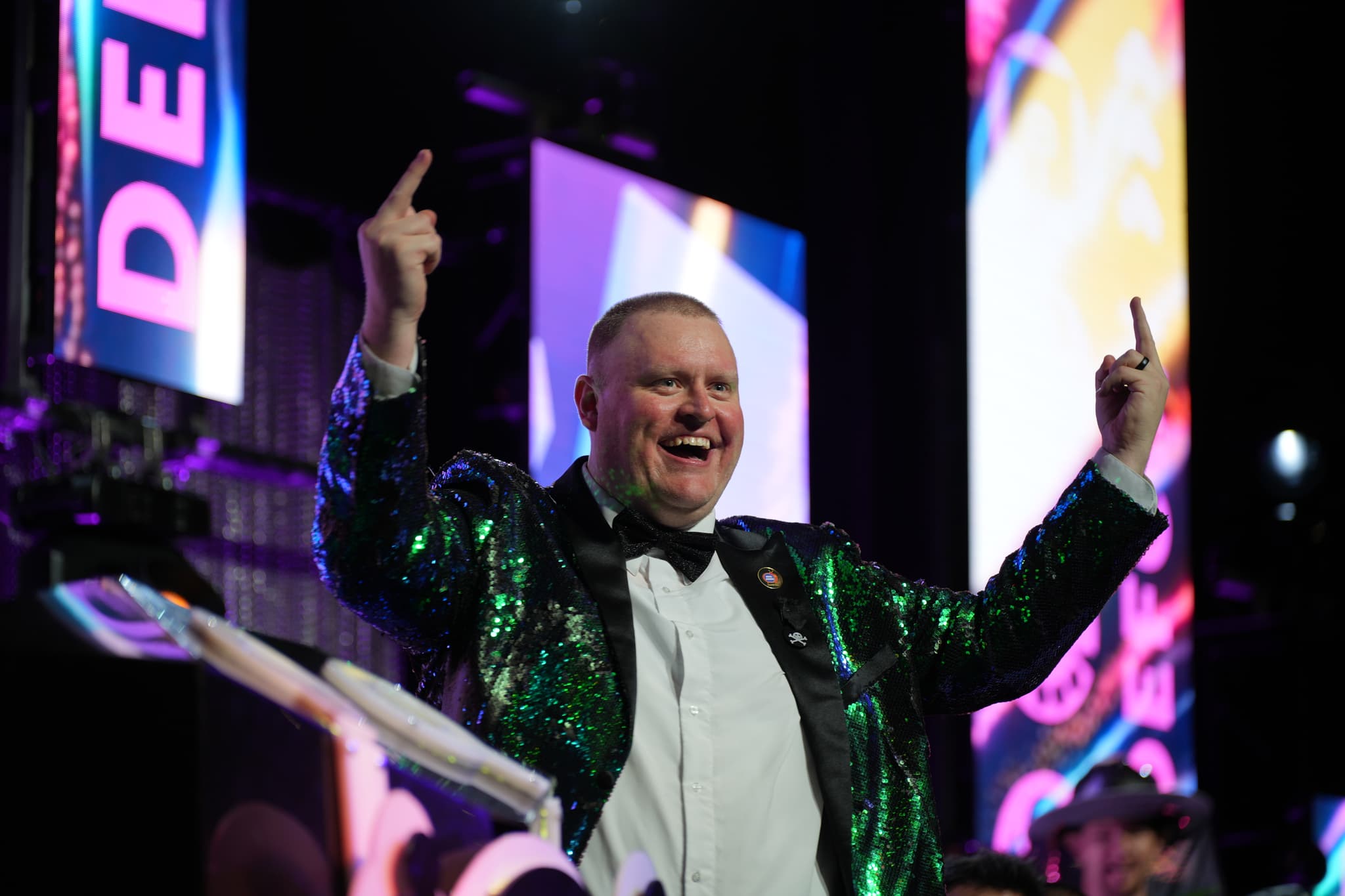 A man in a sparkly green jacket is smiling and raising both hands with his index fingers pointing upwards, standing in front of a colorful, illuminated background