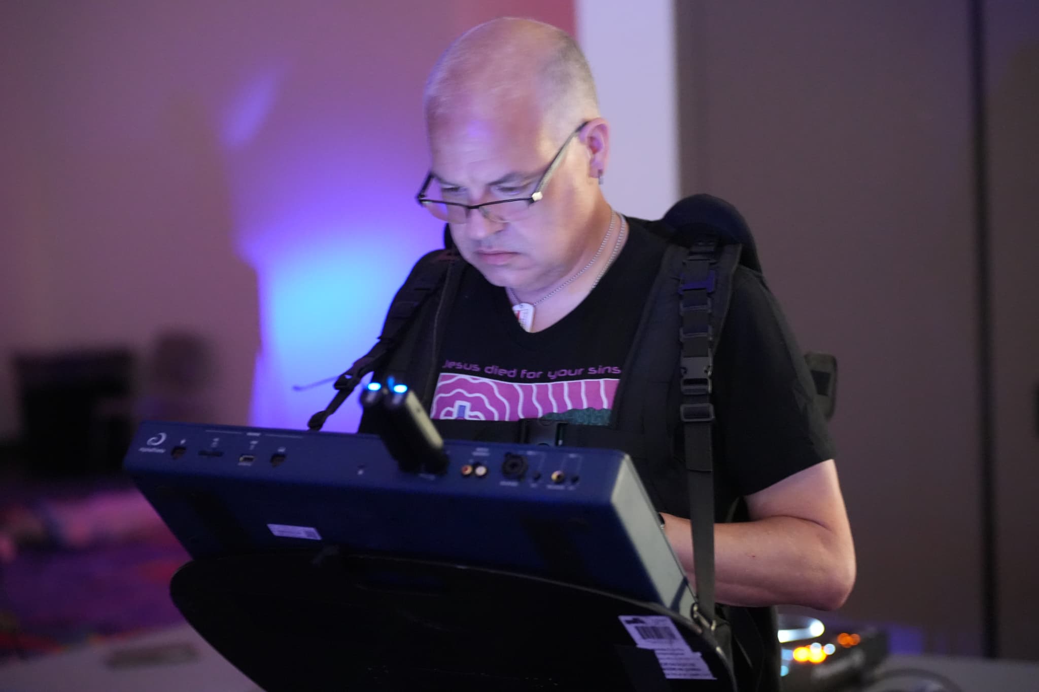 A person wearing glasses and a black t-shirt is focused on operating a piece of electronic equipment, possibly a synthesizer or a control panel, in a dimly lit room with a blue light in the background