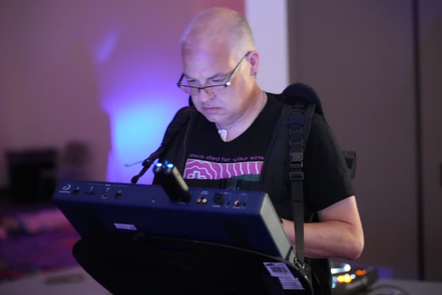 A person wearing glasses and a black t-shirt is focused on operating a piece of electronic equipment, possibly a synthesizer or a control panel, in a dimly lit room with a blue light in the background