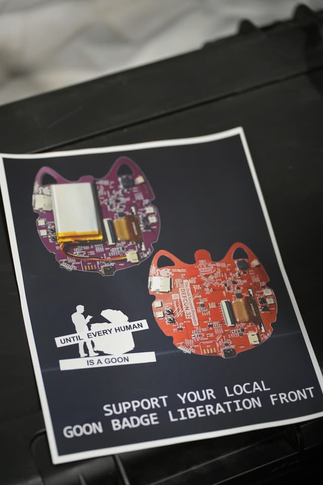 A flyer featuring two electronic circuit boards shaped like cat heads, one purple and one red, with the text SUPPORT YOUR LOCAL GOON BADGE LIBERATION FRONT and an illustration of a person holding a large badge